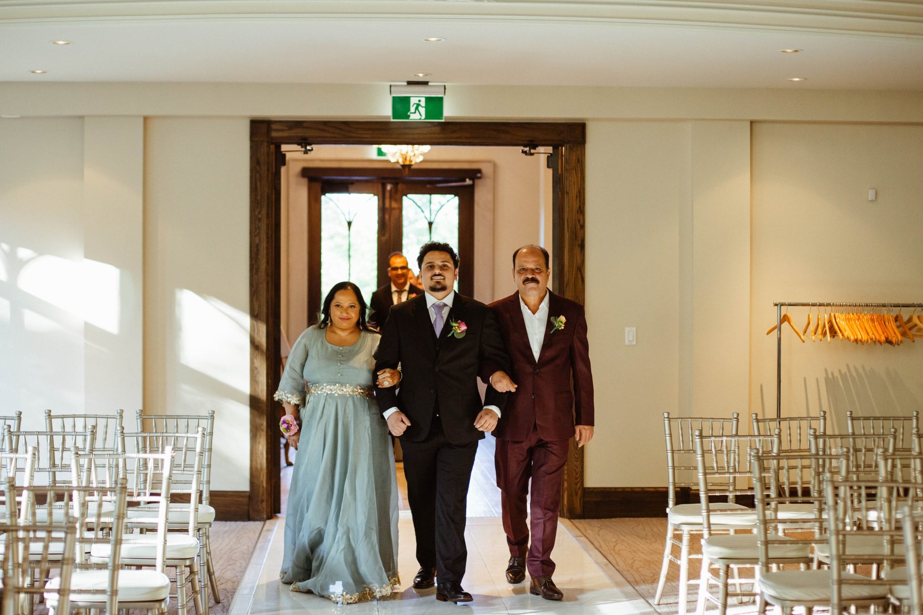 Groom walking down the aisle at Ancaster Mill, arm in arm with his mother and father.