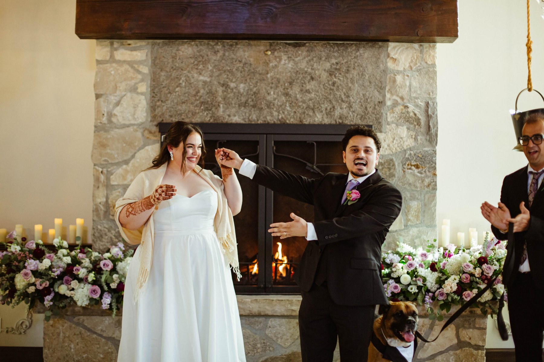 Newlyweds raising their joined hands in celebration in front of the flower-covered fireplace at Ancaster Mill, their dog looking on.
