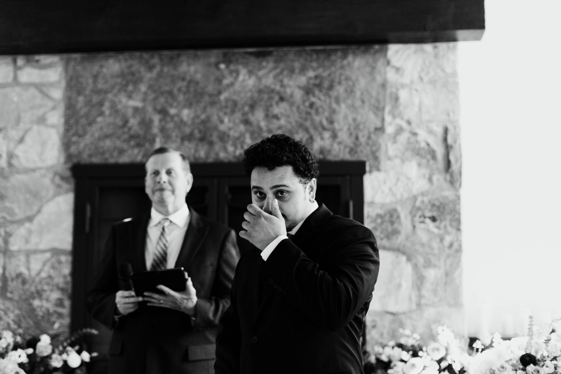 Black and white photo of a groom wiping away tears in front of the stone fireplace at Ancaster Mill as the officiant looks on.