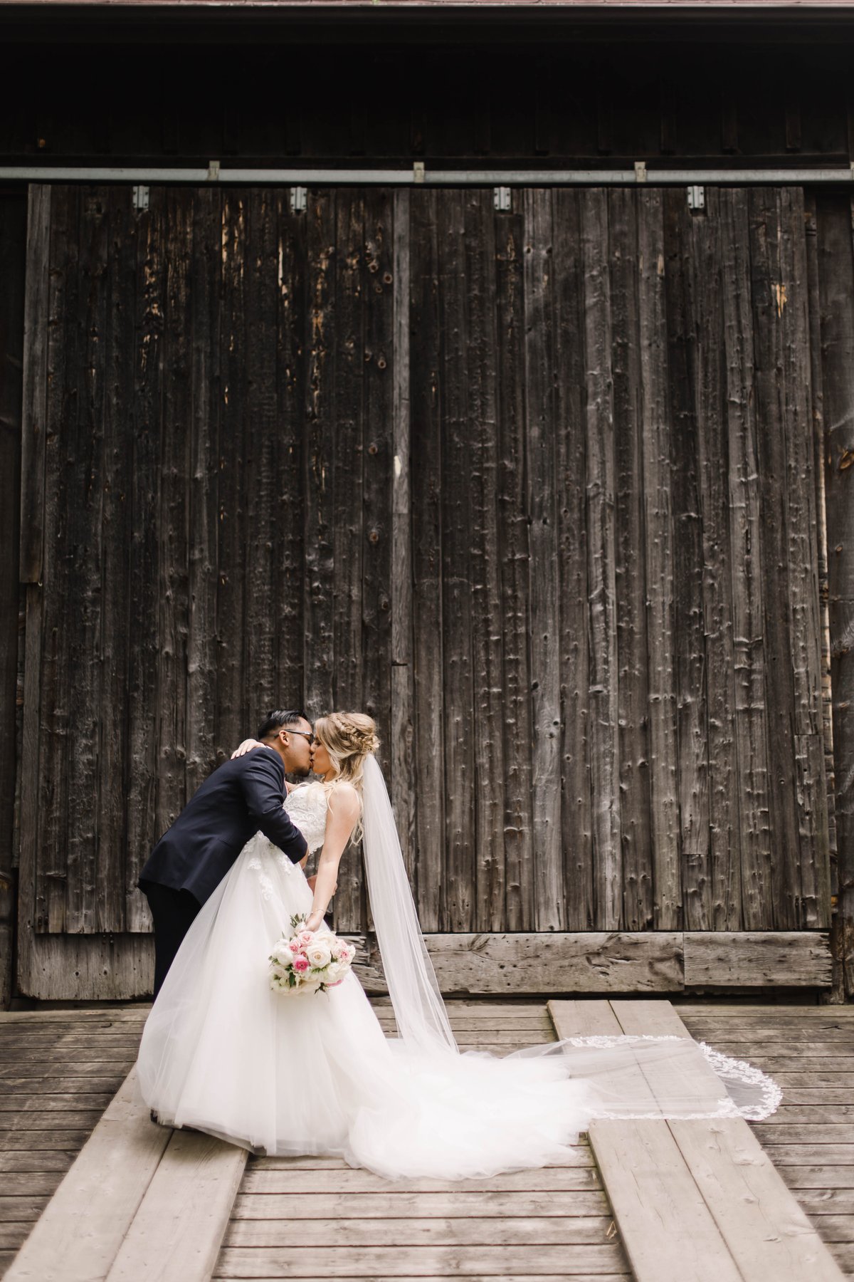 Bride and groom kissing in front of a weathered wooden barn, holding a pink and white bouquet.