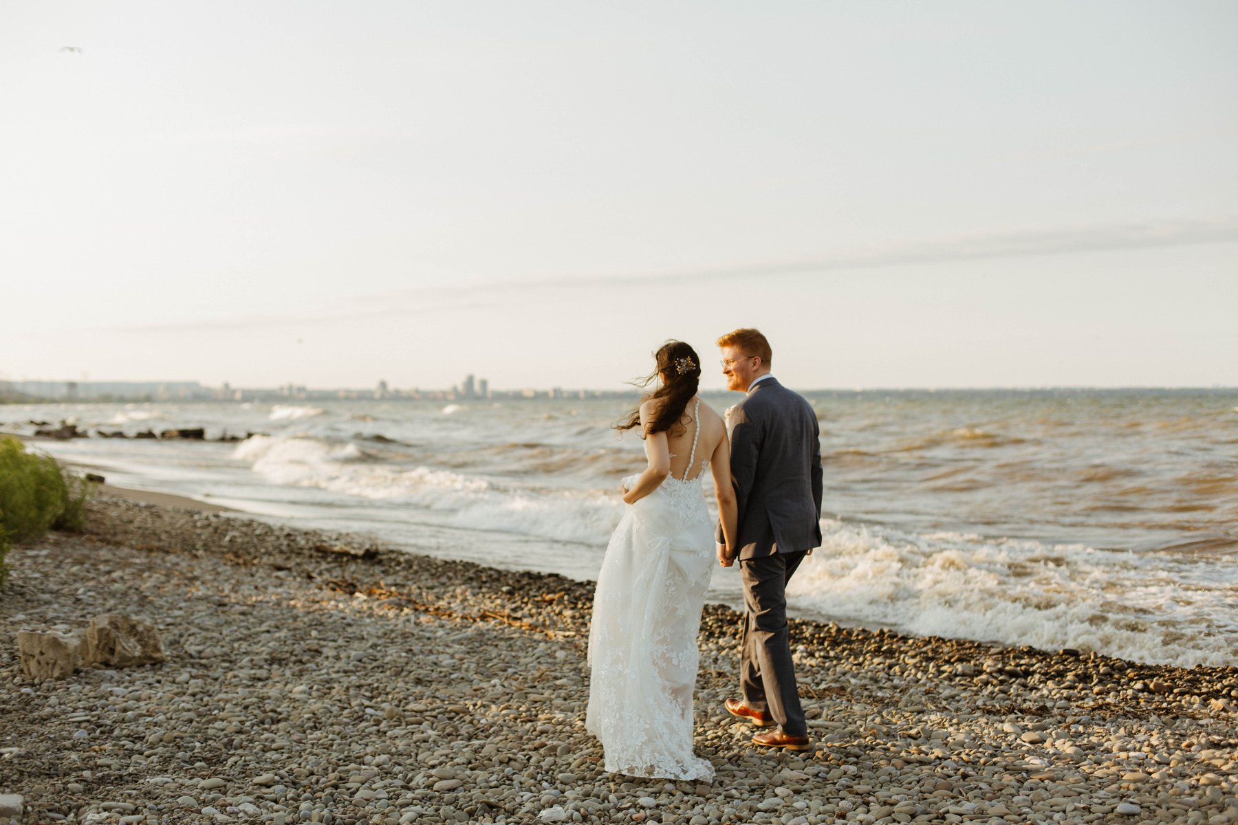 Bride and groom walking hand in hand along a pebble shoreline with a distant skyline.