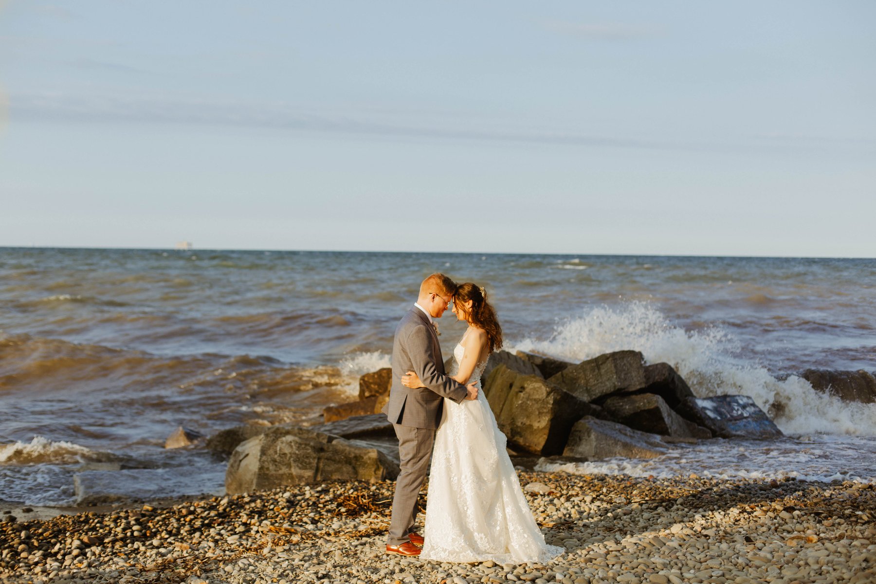 Bride and groom embracing on a breakwater as a wave crashes beside them.