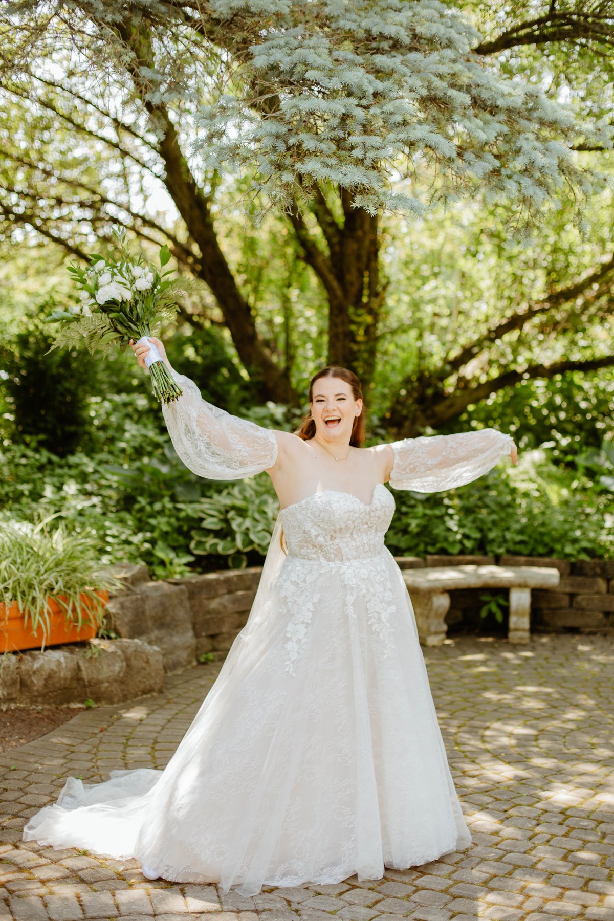 Bride laughing with arms raised, holding her bouquet, under a large tree.
