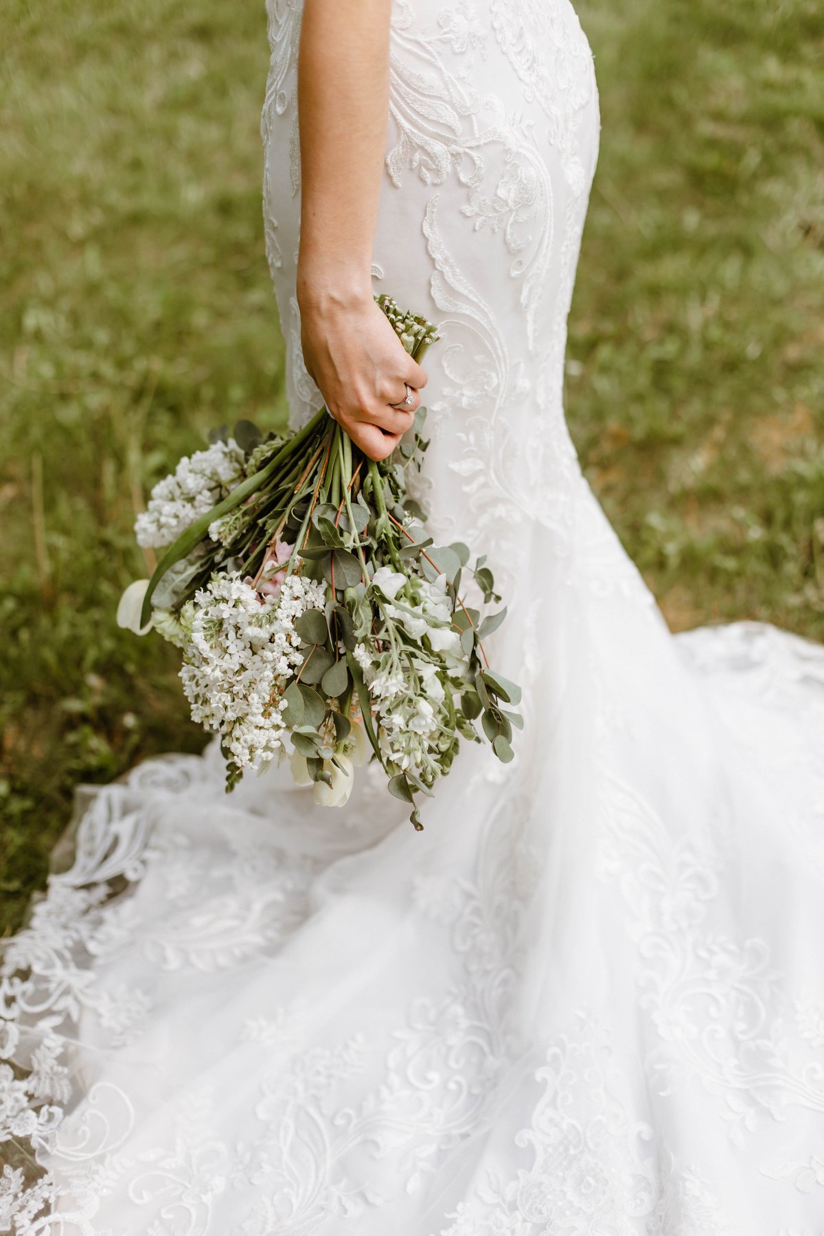 Close-up detail of a bride's bouquet and lace dress train in the grass.