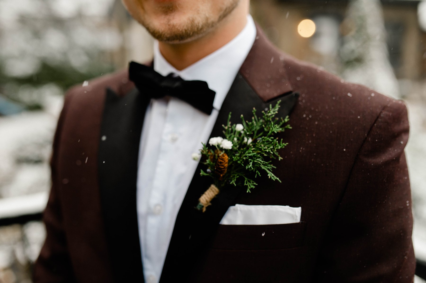 Close-up of a groom's boutonniere on a burgundy suit as snow falls.