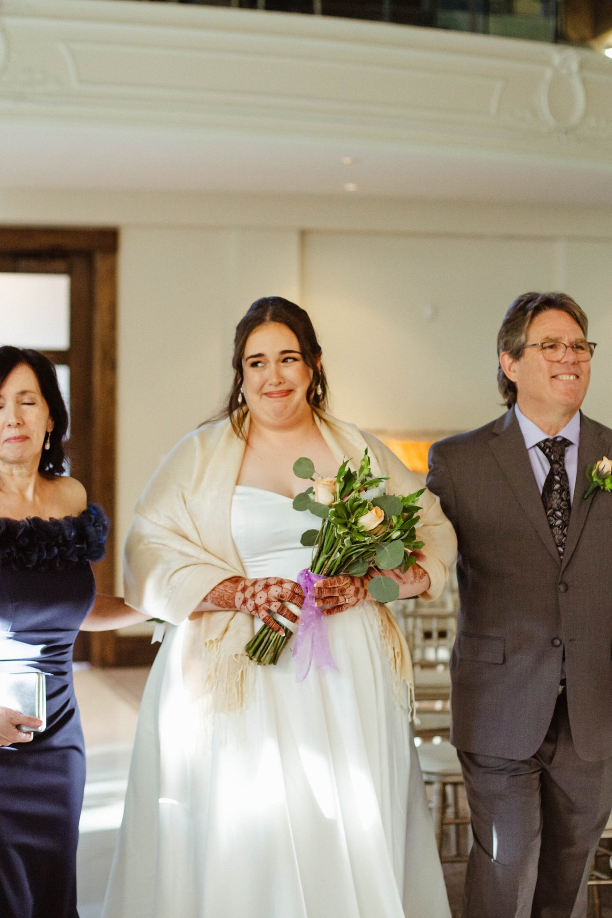 Bride walking down the aisle between her parents, holding her bouquet and smiling.