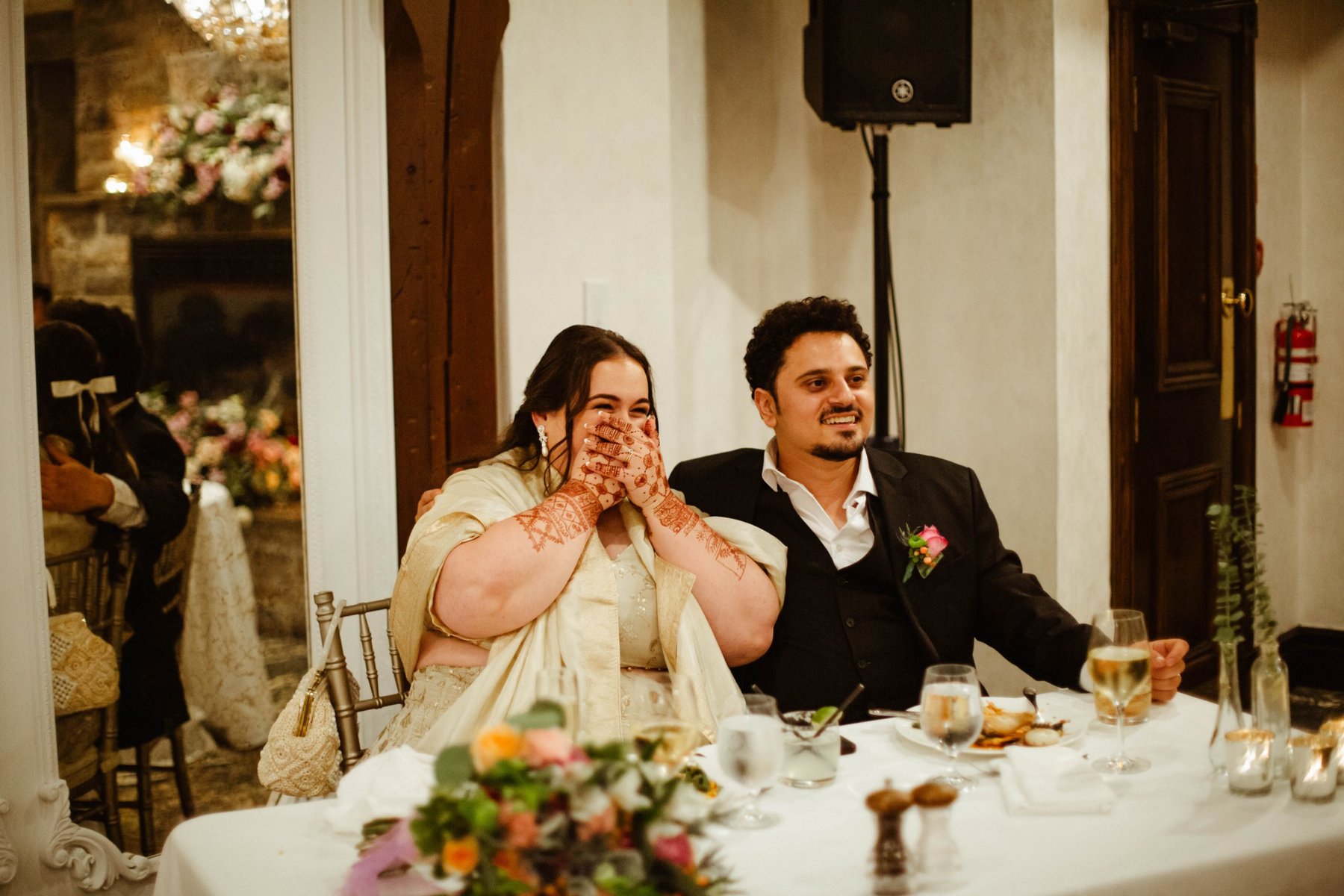 Bride laughing with her hands over her mouth beside her groom during speeches at the reception.