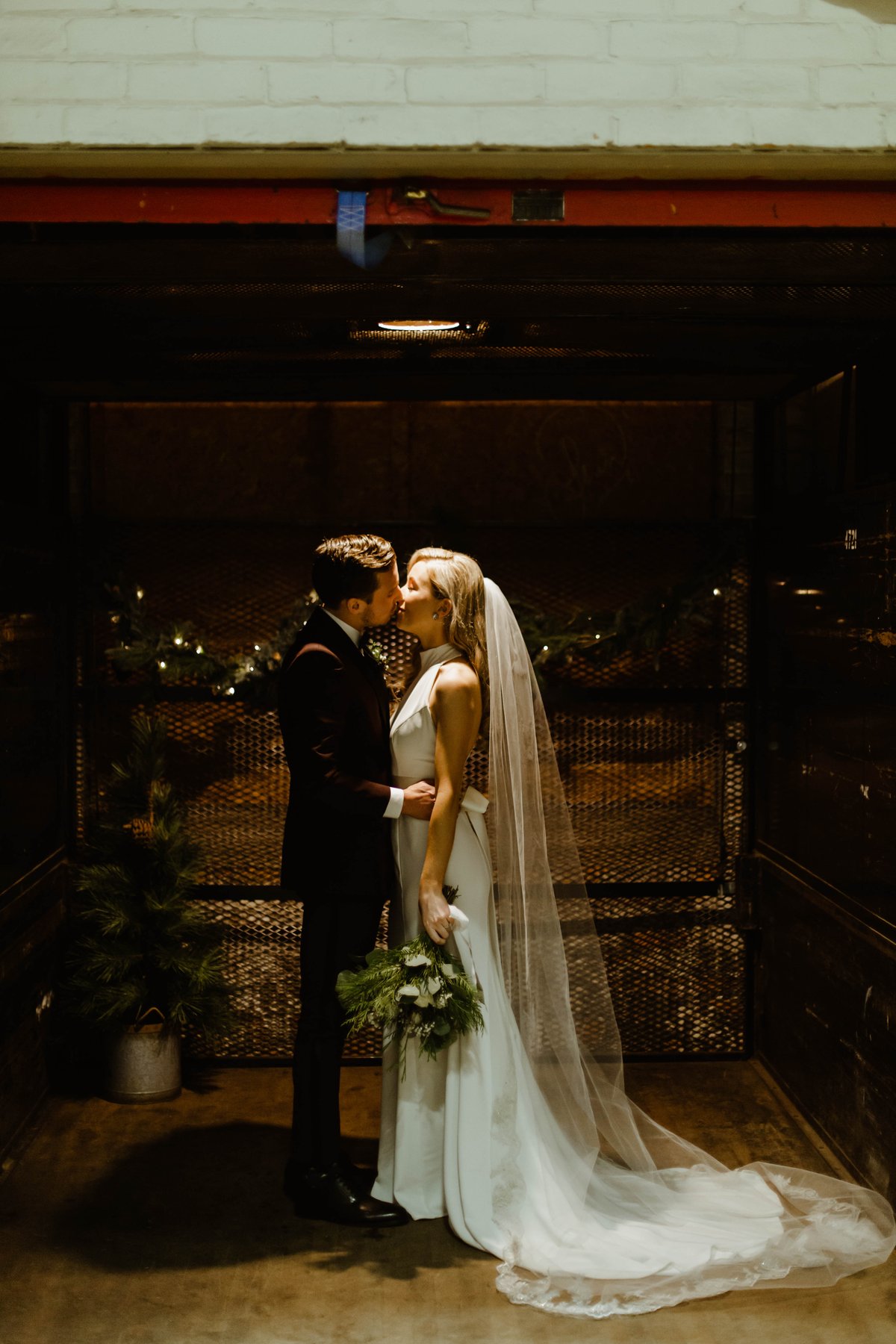 Bride and groom kissing inside a dimly lit freight elevator decorated with greenery.