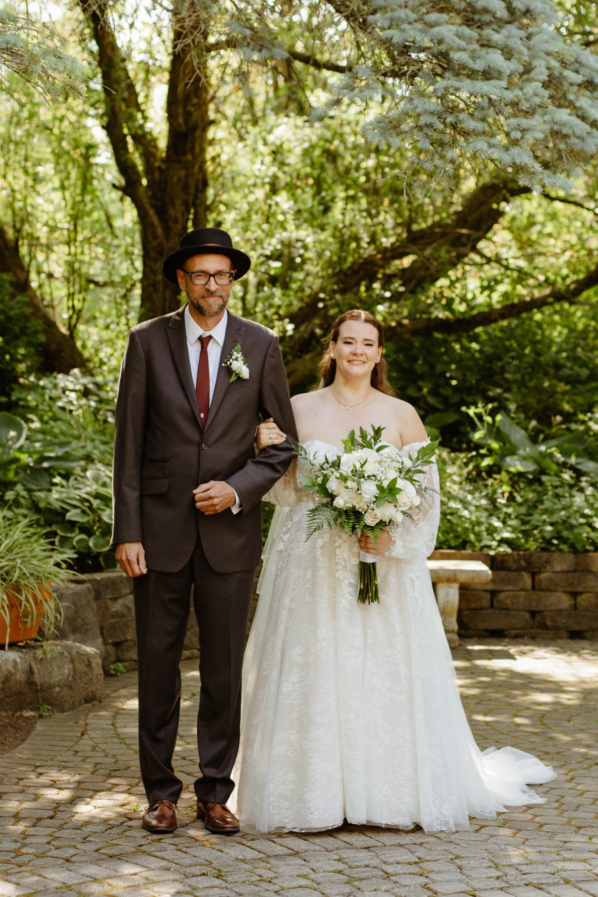 Bride walking arm in arm with her father, both smiling, under trees.
