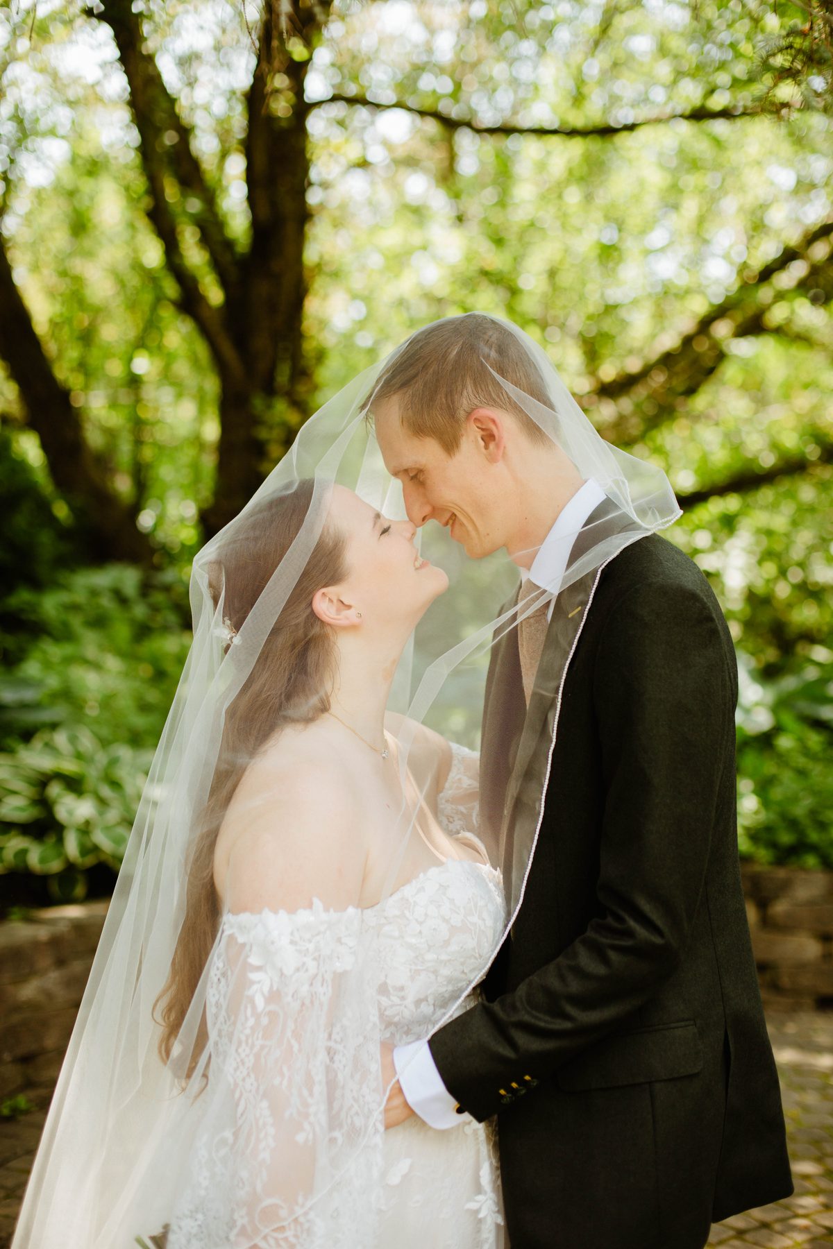 Bride and groom kissing under her veil in a sunlit forest.