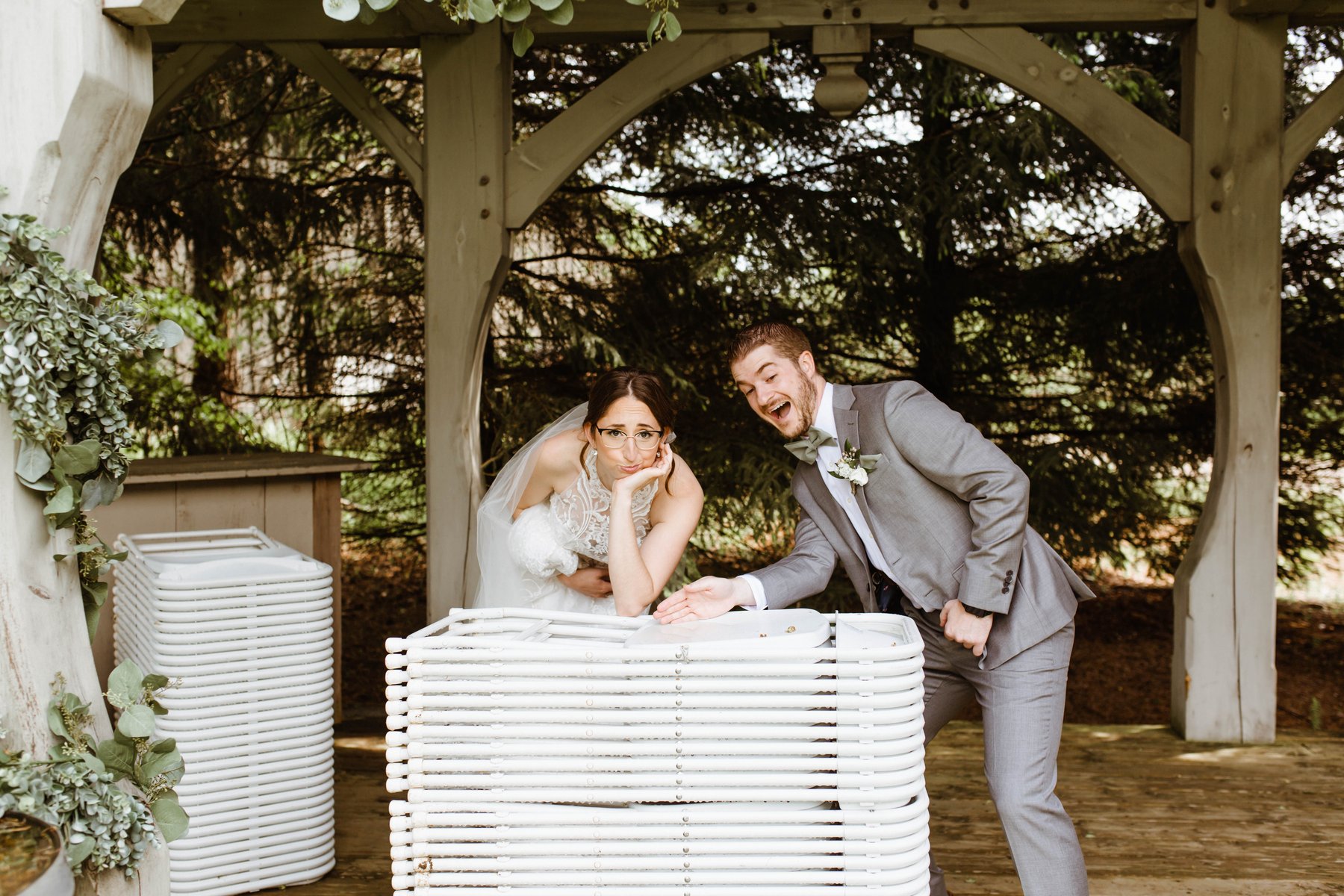Bride and groom making silly faces while leaning on stacked patio furniture at their venue.