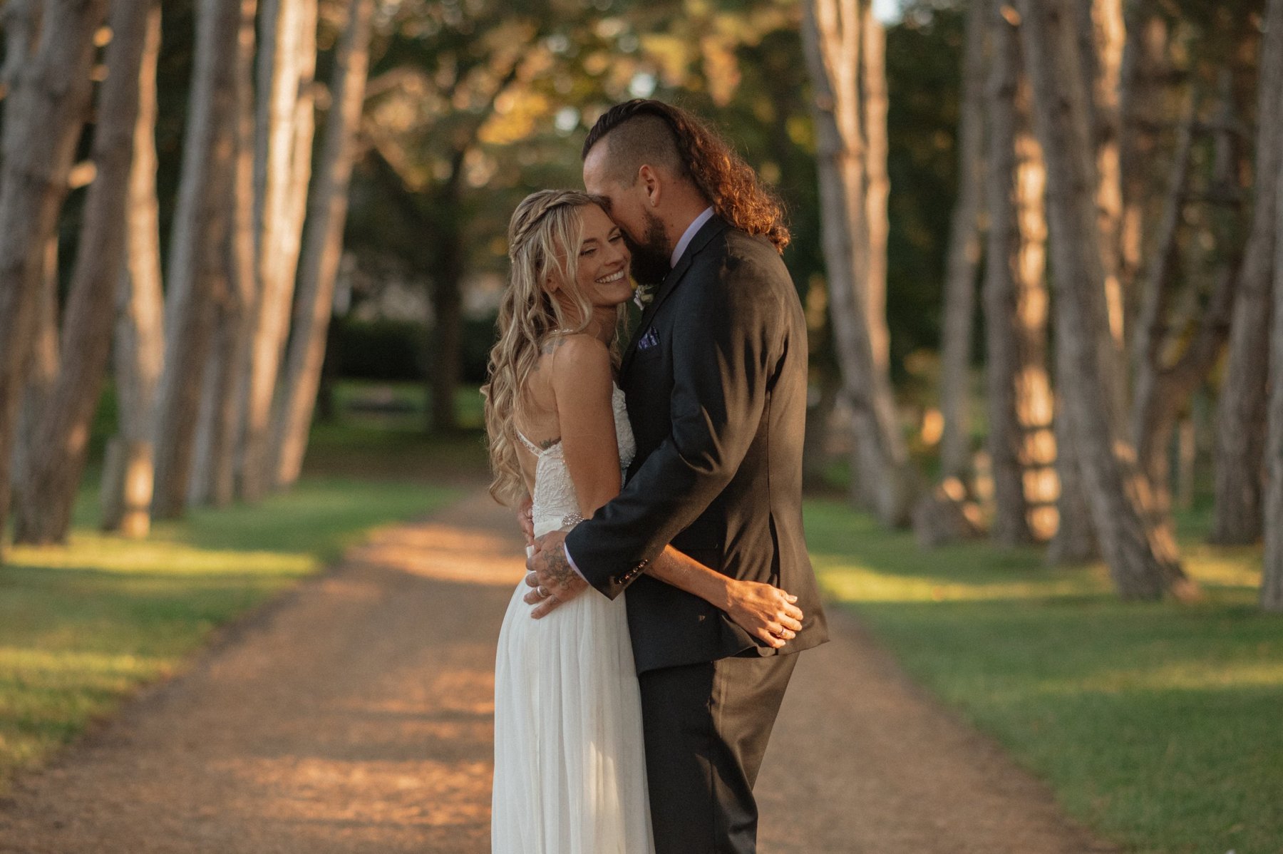 Groom kissing the bride's forehead on a sunlit, tree-lined path at golden hour.