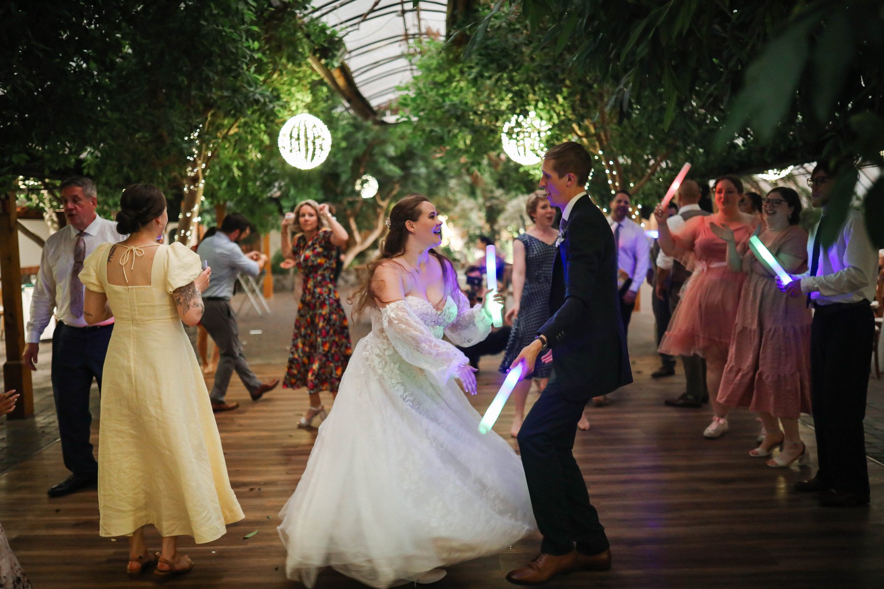 Bride and groom dancing with glow sticks surrounded by cheering guests in a greenhouse reception.