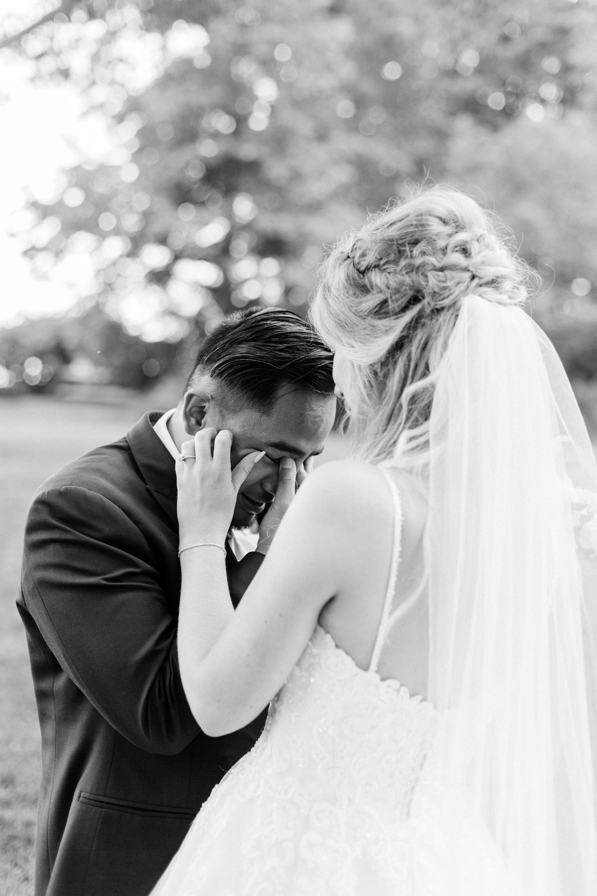 Black and white close photo of a groom wiping away tears as his bride comforts him.