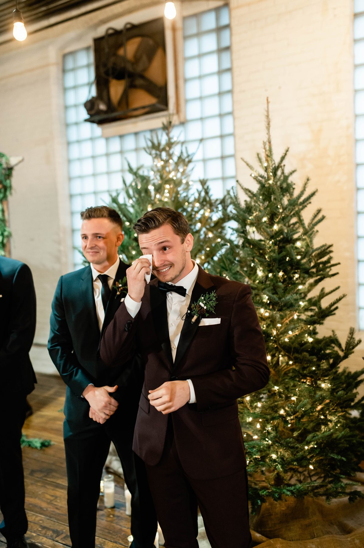 Groom wiping his eyes in front of lit Christmas trees while his groomsman looks on.