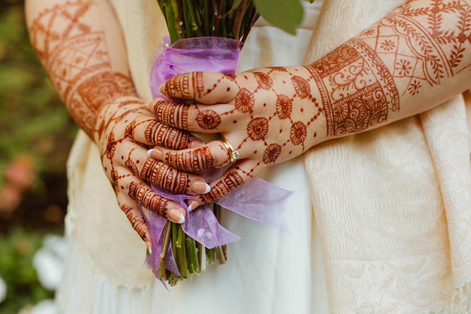 Close-up of a bride's henna-painted hands holding her bouquet, wrapped in purple ribbon.