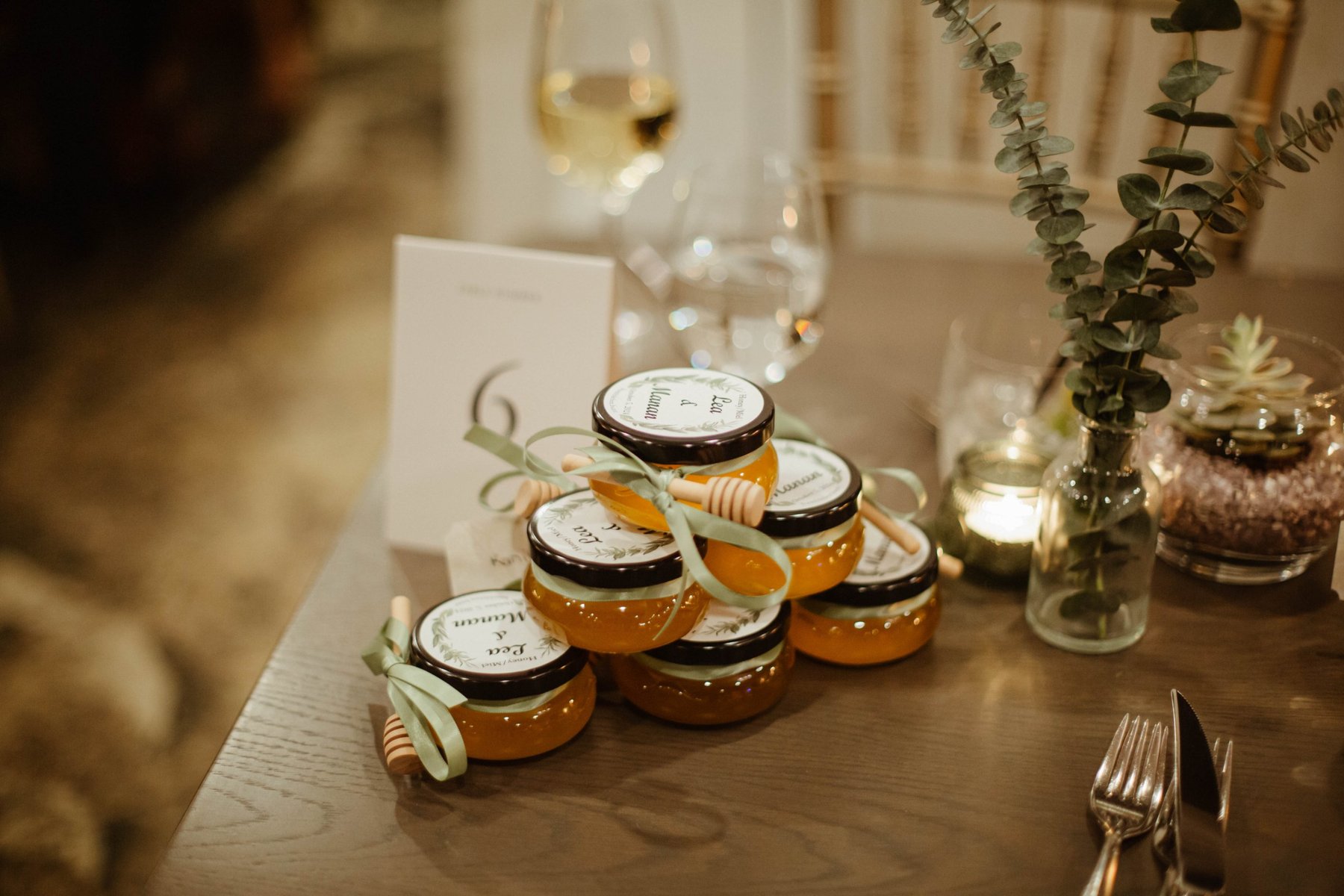 A stack of personalized honey jar wedding favors on a candlelit reception table.