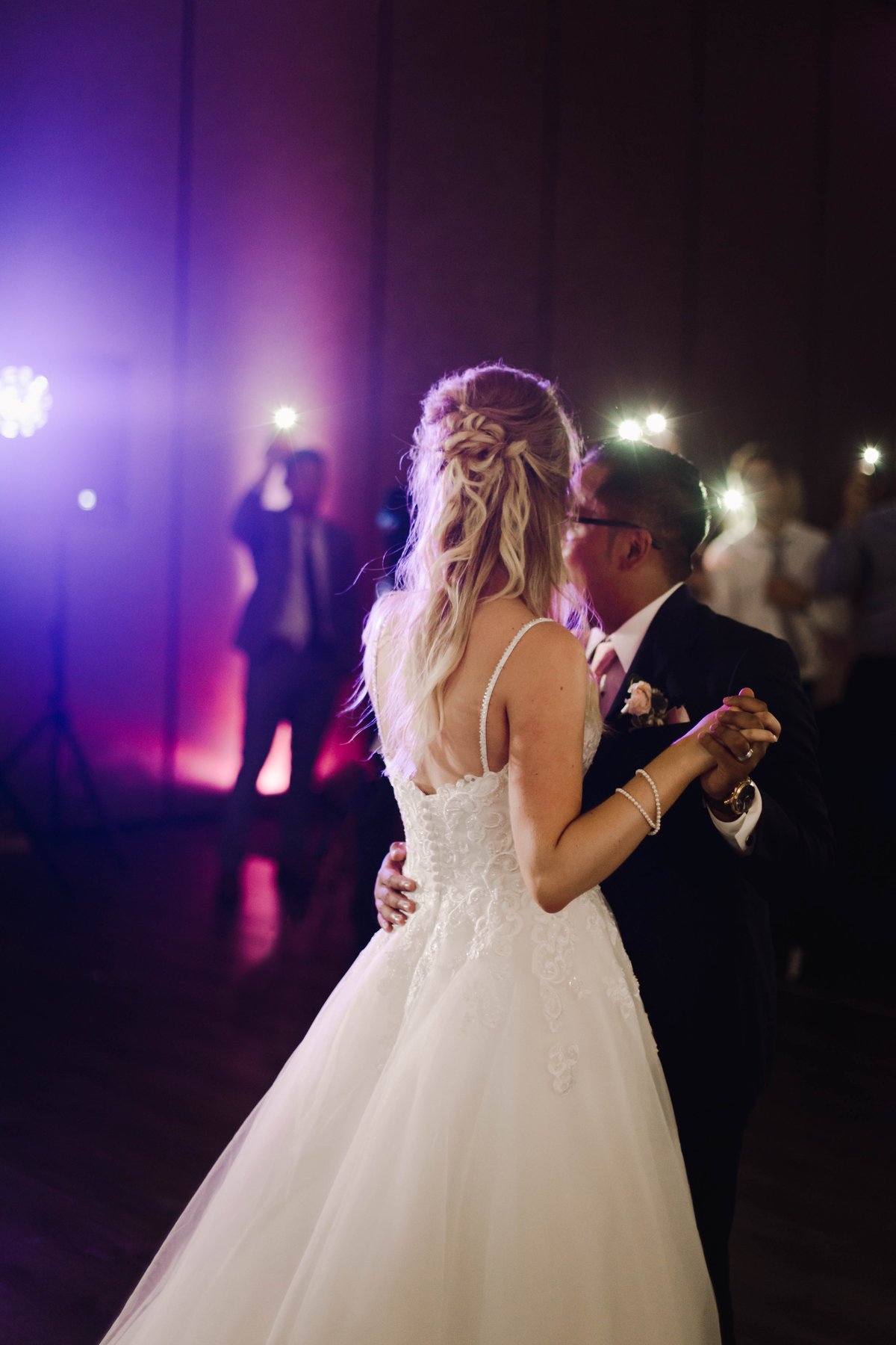 Bride and groom dancing together under purple reception lighting as guests take photos.