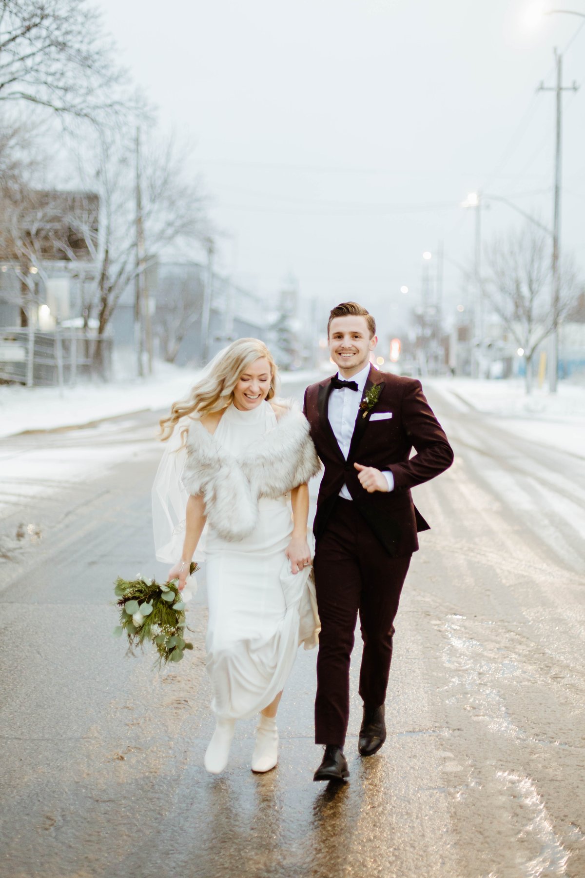 Bride and groom laughing while walking down a snowy street together.
