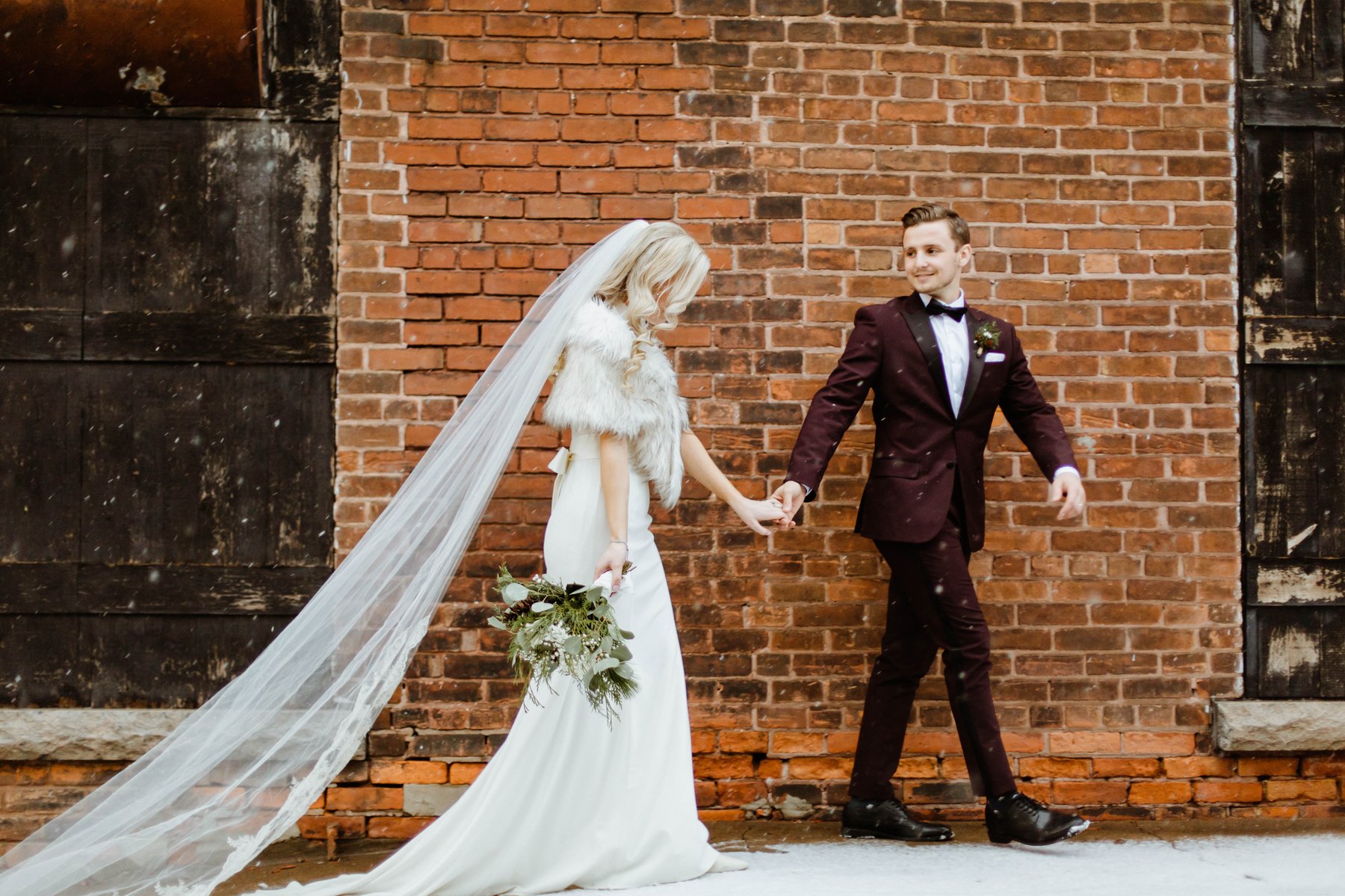 Bride and groom holding hands and walking past a brick wall as snow falls.
