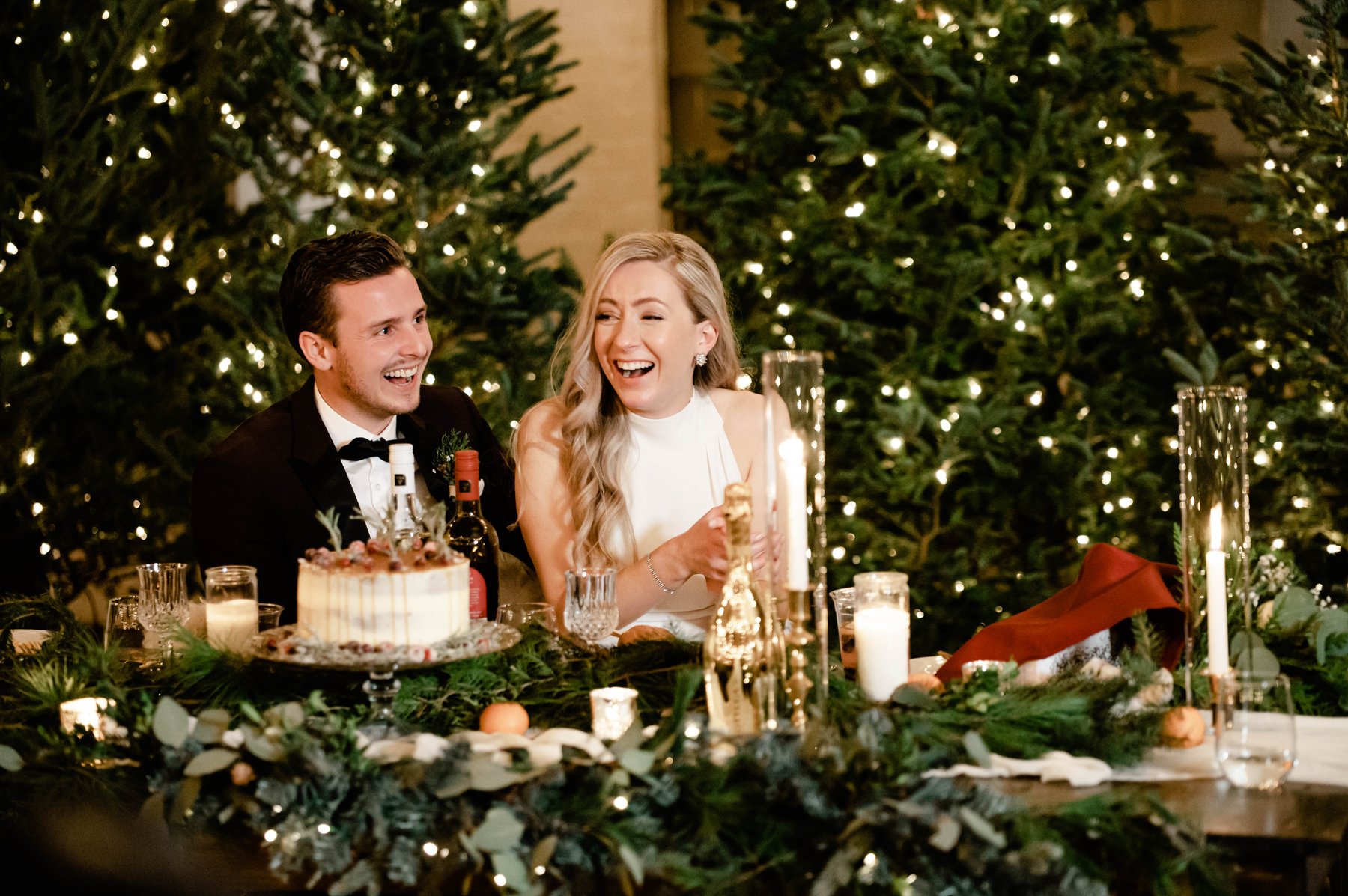 Bride and groom laughing at their sweetheart table surrounded by lit Christmas trees.
