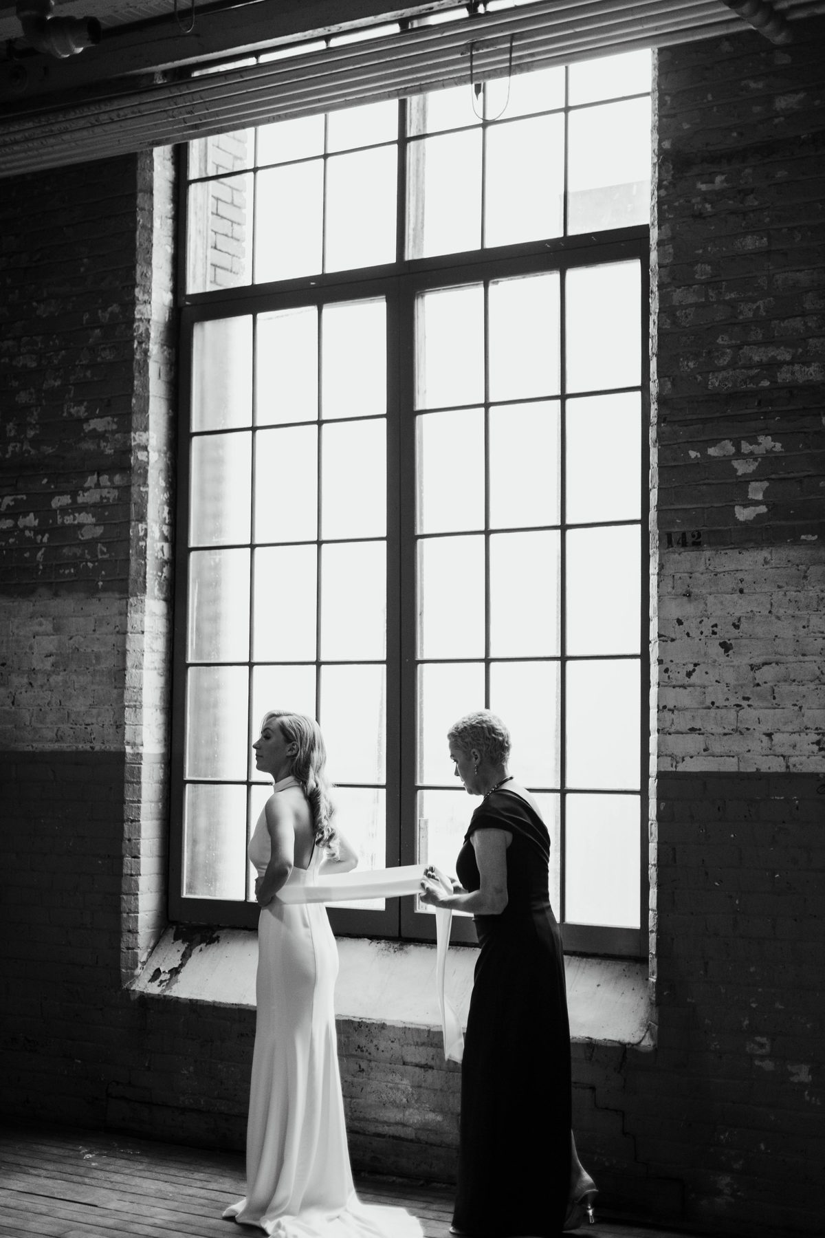 Bride having her dress adjusted in front of a large factory window before the ceremony.