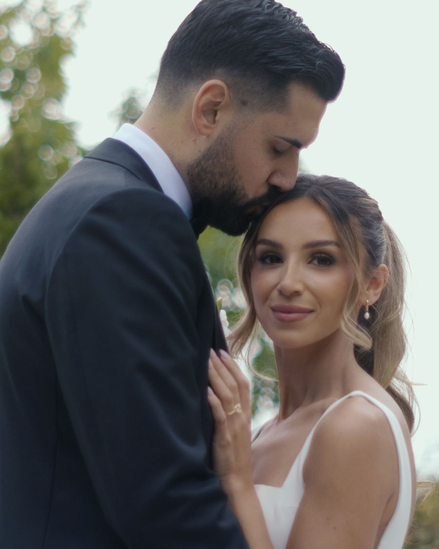 A close, intimate portrait of a groom kissing his bride's forehead outdoors.