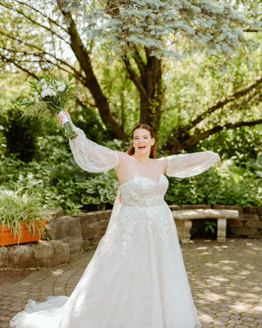 Bride laughing and twirling with her bouquet raised in a garden.