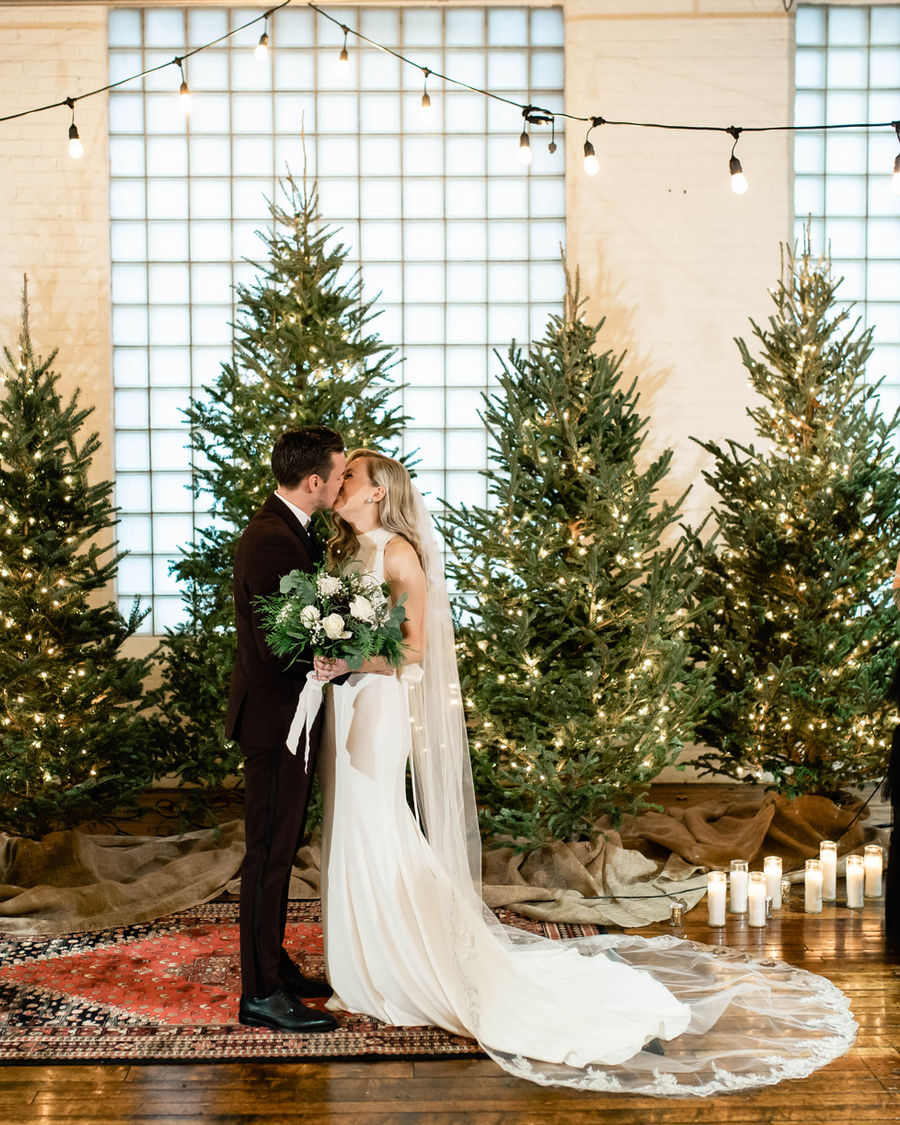 Couple kissing in front of lit Christmas trees at an indoor winter ceremony.
