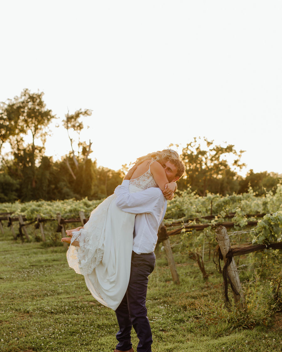 Groom lifting his bride in a hug at golden hour in a vineyard.